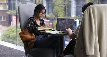 Two students sitting at table next to large glass window working on laptops