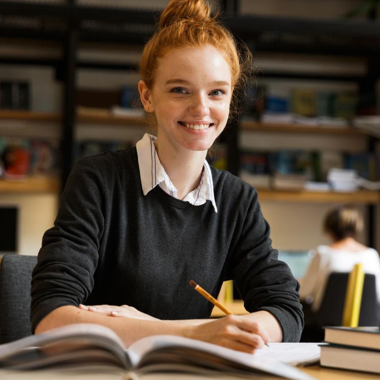 Student sitting at desk writing in an open book and smiling at the camera