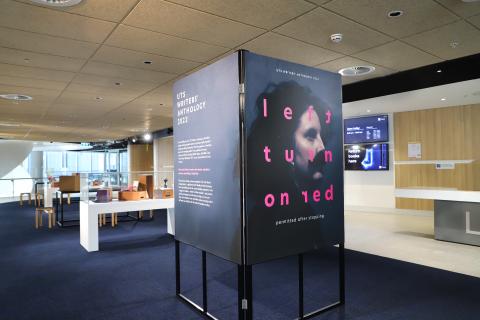 Photograph displays Left turn on Red exhibition setup. A large right angle frame is covered with a large poster with the exhibition title and image of a woman with a moustache