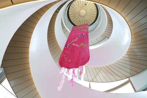 We look up at a vibrant pink fabric tapestry hanging down from the ceiling. Positioned in the middle of a large circular staircase, the installation ends high above our heads, in streamers of transparent dark pink and light pink.